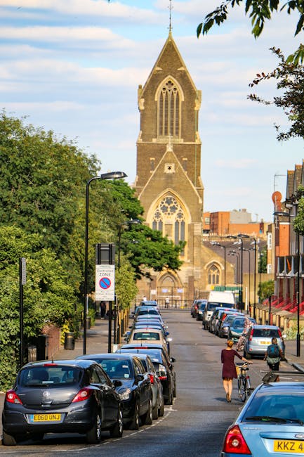 A street scene showing a line of parked cars along a residential road with a woman and a person riding a bicycle visible in the foreground. In the background, there is a historic stone church with a tall, pointed steeple and large arched windows, surrounded by trees and modern buildings. The street is lined with lampposts and signage, including a parking restriction sign. The scene is well-lit with natural daylight, indicating clear weather. This urban setting depicts a typical area where house removals or furniture transport services by Man with Van Newington might operate, with the church serving as a notable local landmark in the context of home relocation and moving logistics.