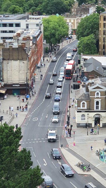 An aerial view of a busy street in Elephant and Castle during daytime, showing multiple parked and moving vehicles including cars and buses, with a narrow lane dedicated to parking along the pavement. Pedestrians are walking along the sidewalks, while some are entering or exiting various buildings, including a white small church-like structure at the corner. Tall green trees line one side of the street, providing shade and greenery. In the background, there are a variety of urban buildings, some with brick façades, showcasing typical London architecture. The scene captures ongoing city activity related to house removals and furniture transport, relevant to home relocation services provided by Man with Van Newington, with street conditions suitable for loading and unloading furniture and boxes during a moving process.
