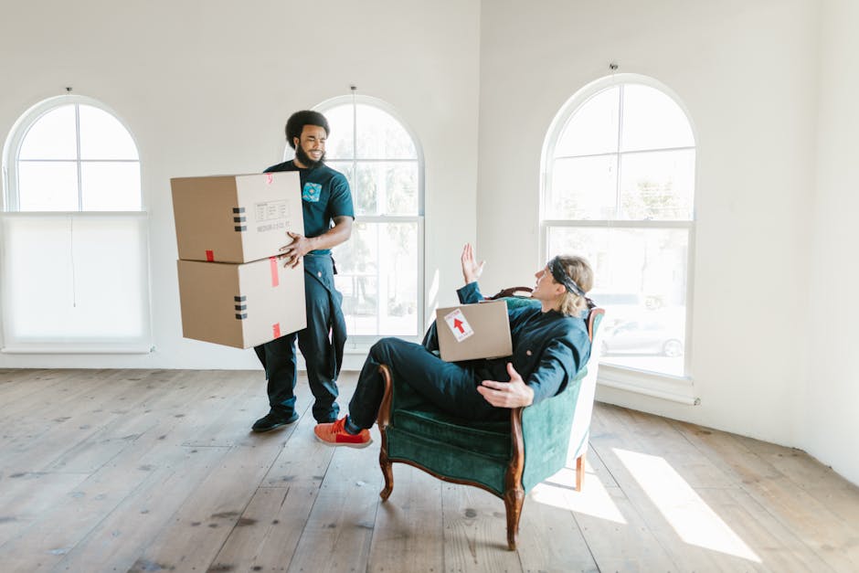 A professional removal team from Man with Van Newington is in the process of a home relocation inside a bright, unfurnished room with white walls and large arched windows, allowing natural daylight to illuminate the space. One mover, standing on a wooden floor, is carrying two medium-sized cardboard boxes stacked one on top of the other, secured with red and black tape. The boxes have packing labels and are being transported from the doorway towards the sofa. The second mover is seated on a vintage-style green armchair with wooden legs, holding a cardboard box with a white label and red arrow sticker, raising a hand to communicate or signal a pause during the furniture transport or packing and moving process. Behind the seated worker, large windows reveal a view of outdoor greenery and parked cars, highlighting the indoor environment where loading and unloading activities for house removals are taking place. The environment suggests a professional and organized approach to moving, with an emphasis on careful handling of household items.