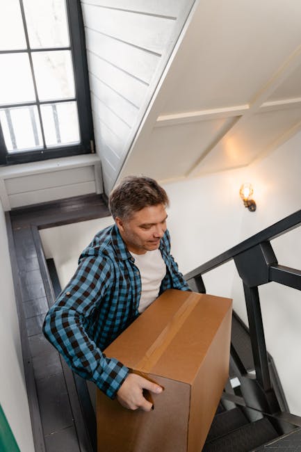 A man with short, dark hair wearing a blue and black checked shirt and light trousers is carrying a medium-sized cardboard box with brown packing tape along the top seam, descending a narrow staircase