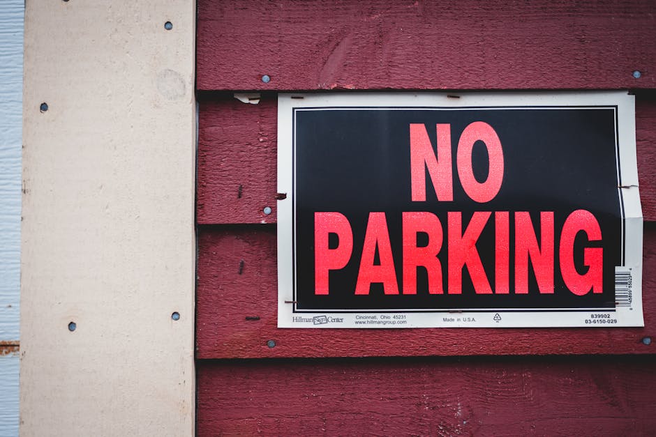A close-up photograph of a rectangular 'No Parking' sign mounted on the exterior red wooden wall of a building. The sign has a black background with bold, uppercase red letters spelling 'NO PARKING'. It is secured with visible metal clips on each corner. To the left of the sign, part of a light-colored beige wall with bolts is visible, contrasting with the textured red wooden siding. The ground below appears to be pavement suitable for loading and unloading activities. This setting suggests a designated area where loading or moving vehicles, such as those used by Man with Van Newington, may access the property for home relocation or furniture transport, particularly relevant in the context of complying with local moving regulations and permits.