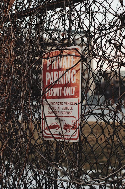 A close-up photograph of a rectangular 'No Parking' sign mounted on the exterior red wooden wall of a building. The sign has a black background with bold, uppercase red letters spelling 'NO PARKING'. It is secured with visible metal clips on each corner. To the left of the sign, part of a light-colored beige wall with bolts is visible, contrasting with the textured red wooden siding. The ground below appears to be pavement suitable for loading and unloading activities. This setting suggests a designated area where loading or moving vehicles, such as those used by Man with Van Newington, may access the property for home relocation or furniture transport, particularly relevant in the context of complying with local moving regulations and permits.