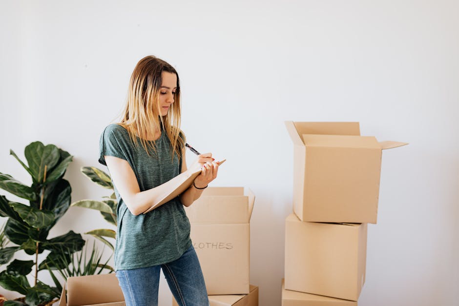 A young woman with long blonde hair, wearing a dark grey t-shirt and blue jeans, is inside a home, standing next to several cardboard moving boxes, some of which are open and others sealed. She is holding a clipboard in one hand and a pen in the other, appearing to be taking notes or checking an inventory list related to packing and furniture transport during a house relocation. The background features a white wall and a large green potted plant, emphasizing a clean, organized interior space. This scene reflects the process of packing and preparing belongings for a move, typical of the services provided by Man with Van Newington, as part of a professional home removals or moving service, with an emphasis on the logistical planning involved in an efficient move.