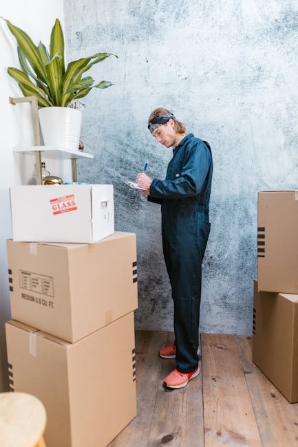 A man wearing a blue jumpsuit, protective goggles, and red shoes is standing inside a room with a textured light blue wall and wooden flooring, surrounded by stacked cardboard moving boxes of varying sizes. Some boxes are sealed with packing tape, with one explicitly labeled 'GLASS' on its side, indicating fragile contents. The man is holding a pen and a clipboard, appearing to take inventory or check off items during a home relocation or packing process. To his left, there is a large potted plant with green leaves on a white shelf attached to the wall. Several boxes are positioned on the floor around him, with some partially opened or sealed, suggesting an ongoing packing or unpacking activity related to house removals. This image illustrates the organization and preparation involved in furniture transport and moving logistics, as part of a professional removal service like Man with Van Newington.