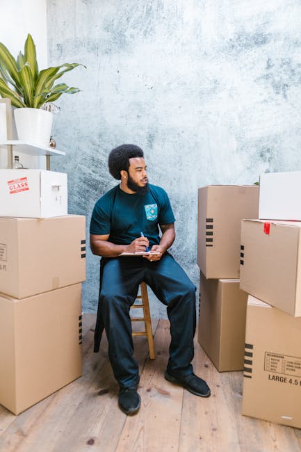 A young woman with long blonde hair, wearing a dark grey t-shirt and blue jeans, is inside a home, standing next to several cardboard moving boxes, some of which are open and others sealed. She is holding a clipboard in one hand and a pen in the other, appearing to be taking notes or checking an inventory list related to packing and furniture transport during a house relocation. The background features a white wall and a large green potted plant, emphasizing a clean, organized interior space. This scene reflects the process of packing and preparing belongings for a move, typical of the services provided by Man with Van Newington, as part of a professional home removals or moving service, with an emphasis on the logistical planning involved in an efficient move.