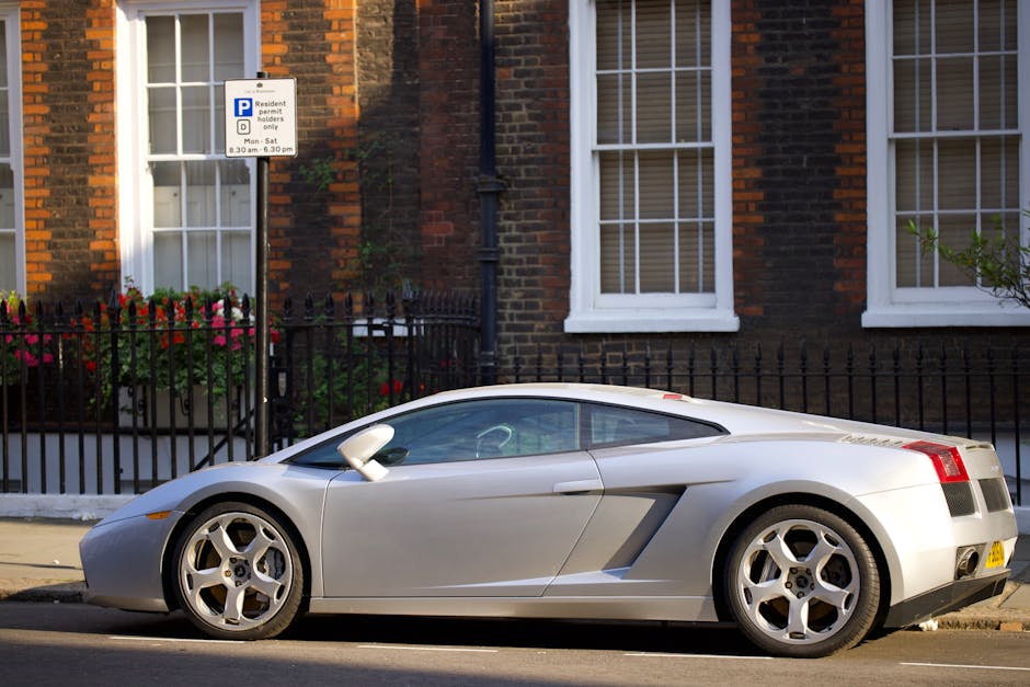 A silver sports car with a sleek, low-profile design is parked on the side of a residential street in Southwark, outside a brick house with white-framed windows. The vehicle's smooth bodywork, alloy wheels, and tinted windows are clearly visible. Behind the car, a black metal fence surrounds a garden with colorful flowers in a white planter box. Above the fence, a parking restriction sign indicates parking permits only between 8:30 am and 6:30 pm, and is attached to a metal pole. The street appears well-lit with natural daylight, emphasizing the car's polished surface and the house's warm brick tones. The scene reflects a typical urban environment where house removals or furniture transport might occur, and highlights the importance of parking permissions during home relocation activities, as managed by local authorities and [COMPANY_NAME].