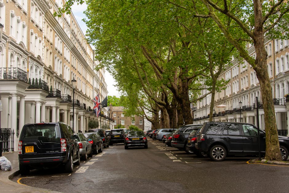 A residential street scene in Southwark with home removal activities by Man with Van Newington. The image shows a narrow street lined with white Victorian-style terraced houses with black wrought iron balconies and flags mounted on some facades. Several parked cars, including a black van on the left and a dark SUV on the right, occupy designated parking spaces along the curb. The pavement is occupied by large, leafy trees with green foliage creating a canopy overhead. Visible inside or near some of the properties are cardboard boxes and wrapped furniture, suggesting packing or moving preparations. The roadway appears wet, reflecting light and adding a shiny texture to the asphalt surface. A vehicle is seen in the process of moving or positioning items during a home relocation or furniture transport operation, while parking suspensions are likely in effect as indicated by the controlled parking environment. This scene captures the typical setting for domestic relocation services in the area, emphasizing both the logistical aspects of moving and the local urban environment.