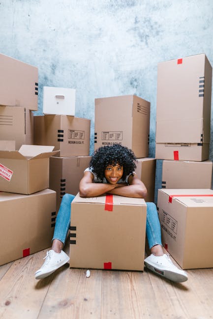 A young woman with curly black hair is seated on the floor inside a room, smiling and leaning on a large cardboard moving box with red packing tape. Surrounding her are numerous additional packed cardboard boxes of various sizes, some with labels or tape, stacked against the light blue textured wall and on the wooden floor. The scene captures a moment during a home relocation or packing process, with boxes arranged for easy loading or transport. The woman is dressed casually in a light-colored top and jeans, and appears ready for a furniture transport or moving process. This setting reflects the packing stage associated with house removals services provided by Man with Van Newington, with the background showing a clean, simple room preparing for a timely move or delivery.