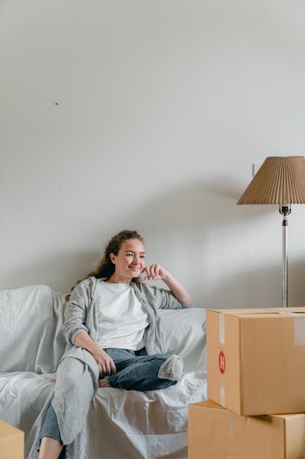 A young woman with light brown, curly hair, wearing a white T-shirt and grey cardigan, is sitting on a white sofa in a room with plain white walls. She has a cheerful expression, smiling and resting her head on her right hand. Beside her on the sofa are four cardboard boxes, two of which are stacked, used for packing belongings during a home relocation. In the background, a tall floor lamp with a beige shade is positioned to the right, illuminating the space with natural indoor lighting. The scene captures a moment during the packing and moving process, with minimal decor and a clean, organized environment, reflecting the preparation for furniture transport by [COMPANY_NAME], a professional removals service specializing in house removals and relocation logistics.