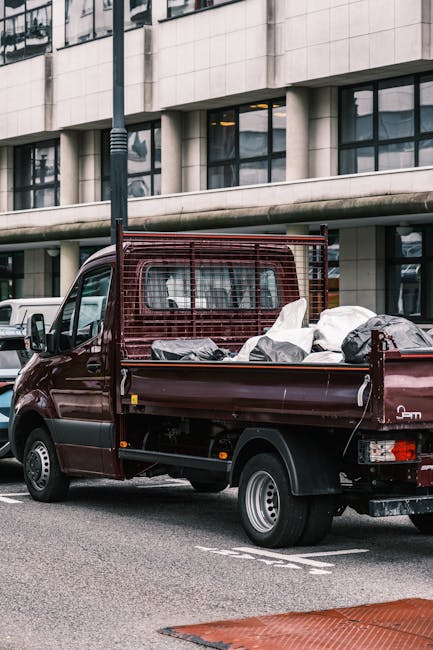 A small burgundy flatbed truck parked on a city street in front of a modern office building with large windows. The truck's bed is loaded with various household items, including white cardboard boxes,