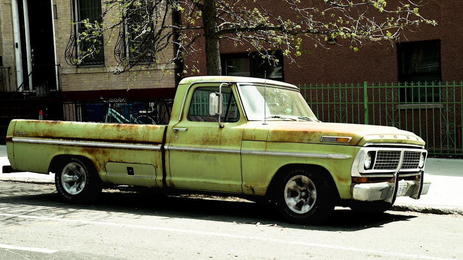 A small burgundy flatbed truck parked on a city street in front of a modern office building with large windows. The truck's bed is loaded with various household items, including white cardboard boxes,